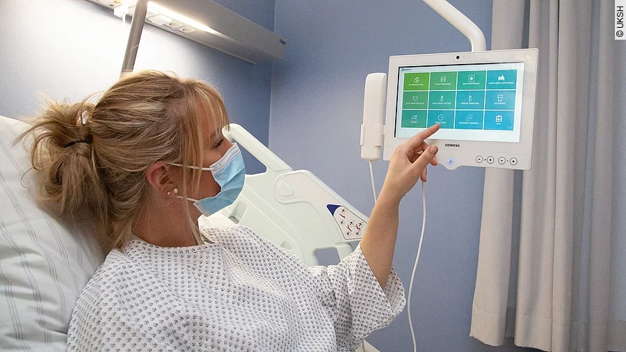 Kommunikationssystem Cliniserve Frau mit Mundschutz im Krankenhausbett tippt auf Monitor.