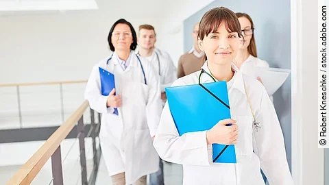 Drei Ärztinnen mit blauen Mappen unterm Arm im Krankenhausflur unterwegs