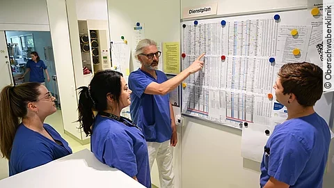 Vier Menschen stehen vor einem Dienstplan in einem Krankenhaus.