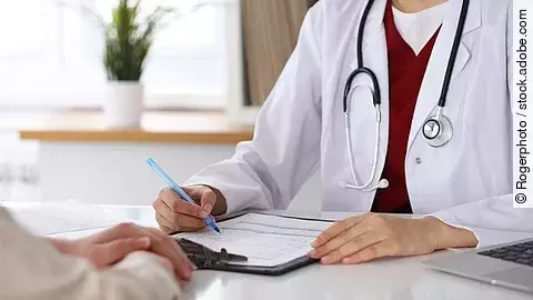 Ärztin füllt Formular aus und sitzt gegenüber eines Patienten.