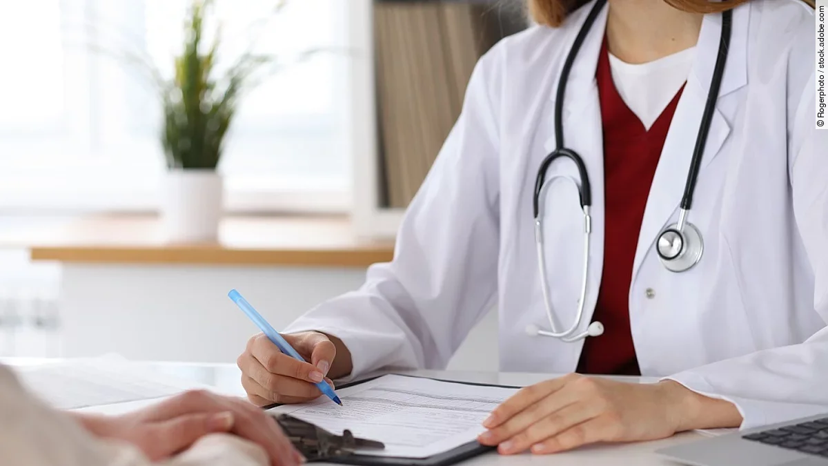 Ärztin füllt Formular aus und sitzt gegenüber eines Patienten.