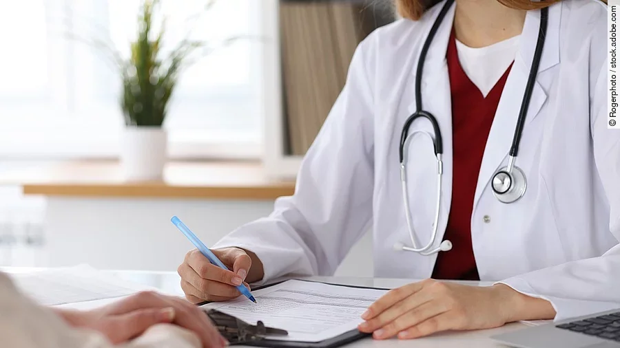 Ärztin füllt Formular aus und sitzt gegenüber eines Patienten.