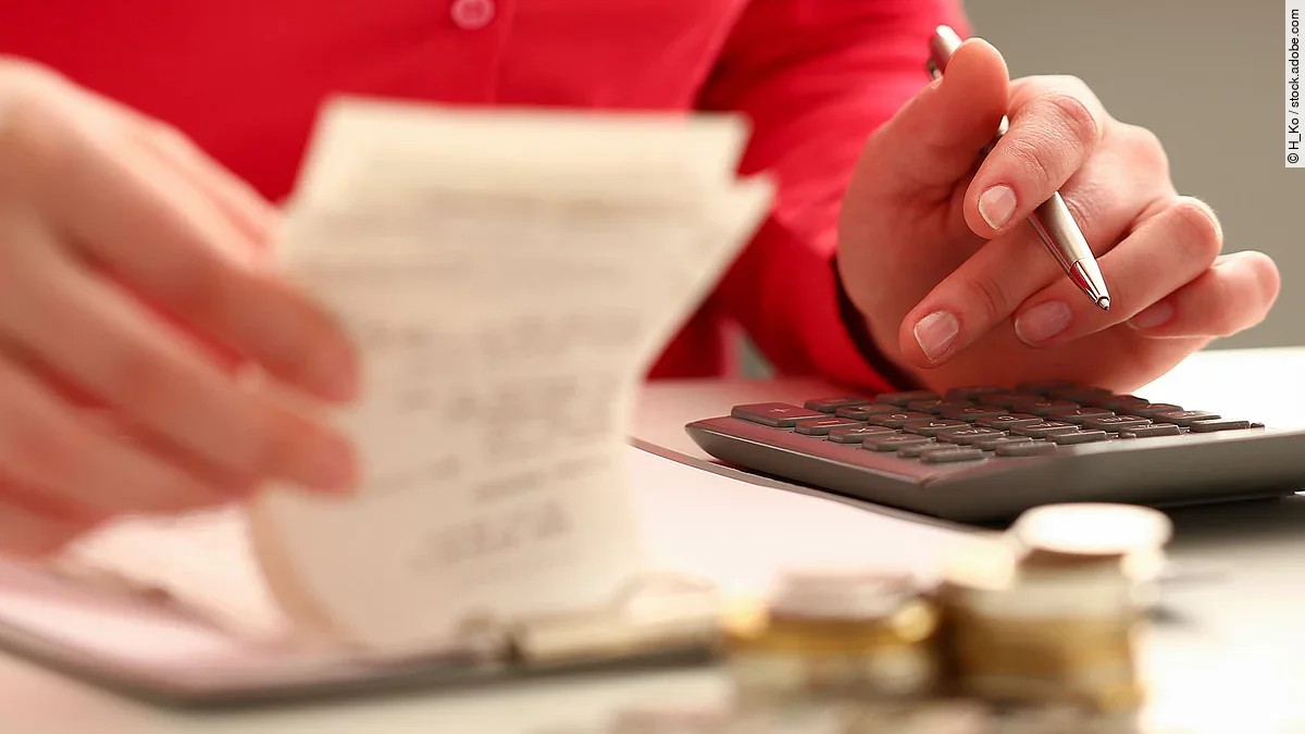Person sitzt an einem Tisch und bearbeitet Rechnungen. Auf dem Tisch steht ein Sparschwein und ein Stapel Münzen.