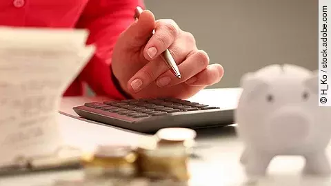 Person sitzt an einem Tisch und bearbeitet Rechnungen. Auf dem Tisch steht ein Sparschwein und ein Stapel Münzen.