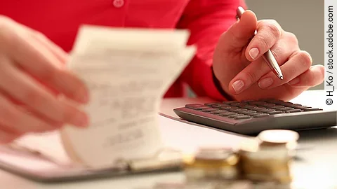 Person sitzt an einem Tisch und bearbeitet Rechnungen. Auf dem Tisch steht ein Sparschwein und ein Stapel Münzen.