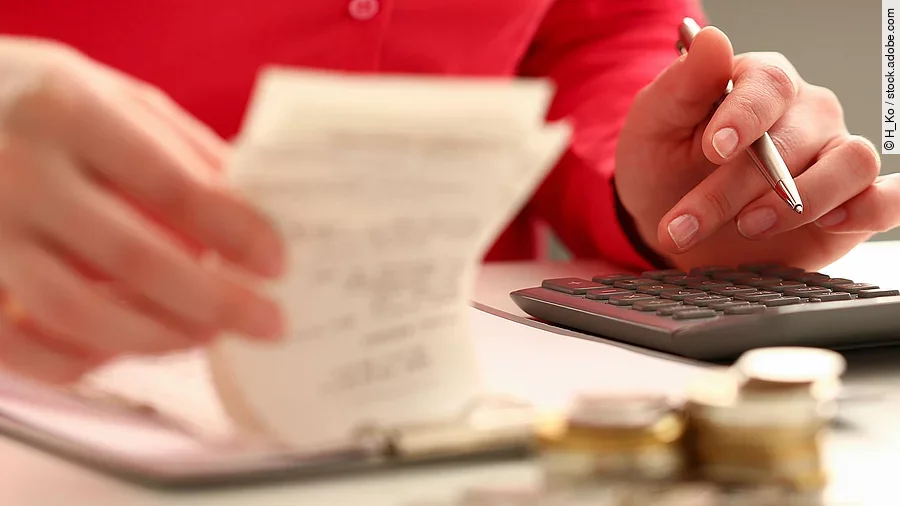 Person sitzt an einem Tisch und bearbeitet Rechnungen. Auf dem Tisch steht ein Sparschwein und ein Stapel Münzen.