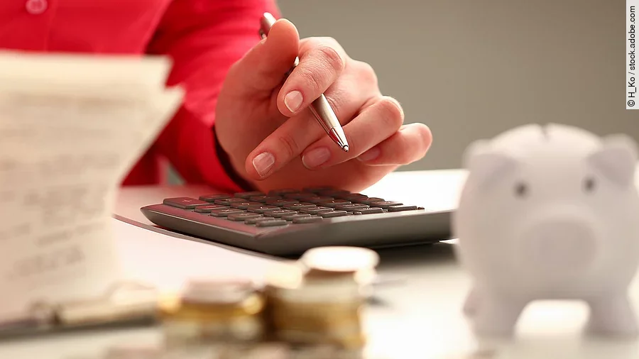 Person sitzt an einem Tisch und bearbeitet Rechnungen. Auf dem Tisch steht ein Sparschwein und ein Stapel Münzen.