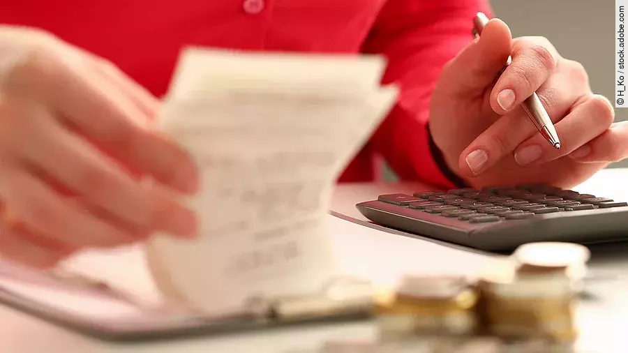 Person sitzt an einem Tisch und bearbeitet Rechnungen. Auf dem Tisch steht ein Sparschwein und ein Stapel Münzen.
