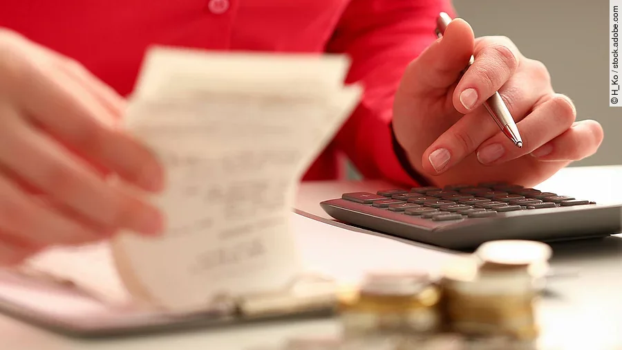 Rechnung Person sitzt an einem Tisch und bearbeitet Rechnungen. Auf dem Tisch steht ein Sparschwein und ein Stapel Münzen.