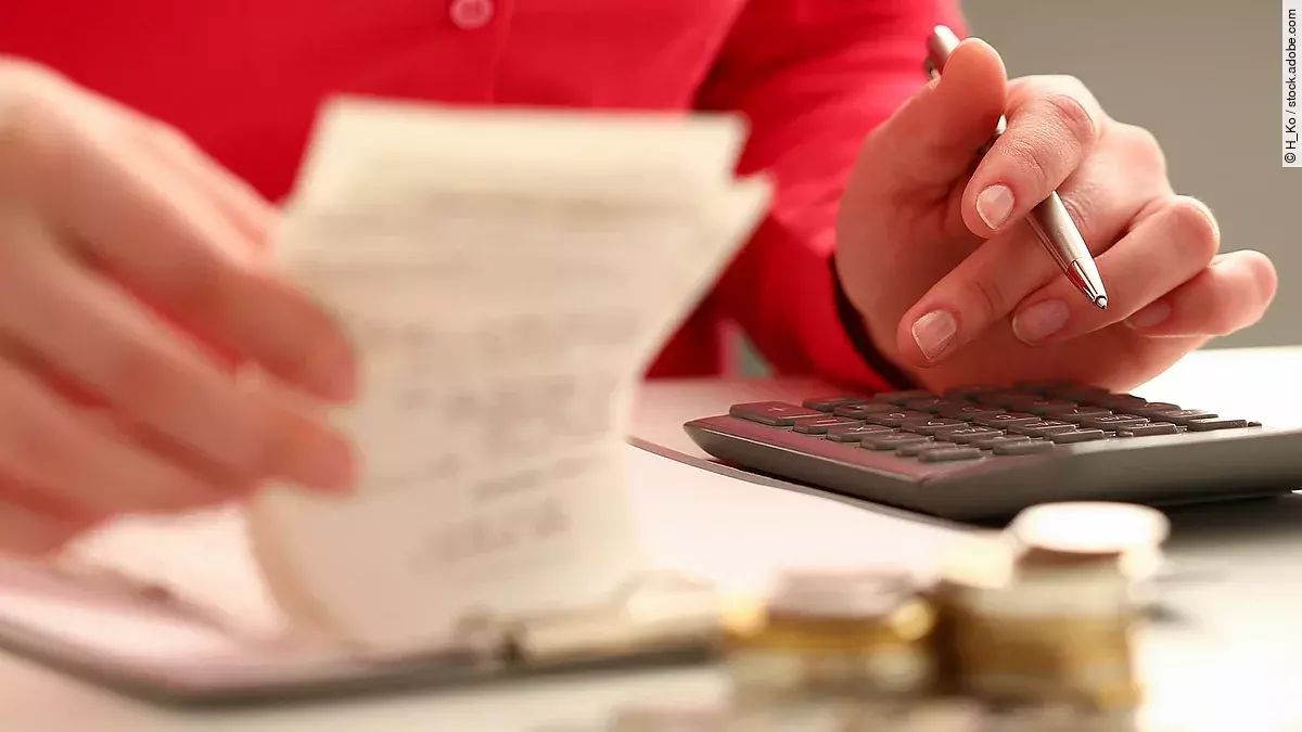Rechnung Person sitzt an einem Tisch und bearbeitet Rechnungen. Auf dem Tisch steht ein Sparschwein und ein Stapel Münzen.