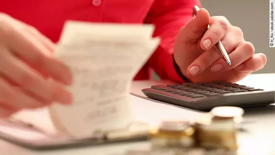 Person sitzt an einem Tisch und bearbeitet Rechnungen. Auf dem Tisch steht ein Sparschwein und ein Stapel Münzen.
