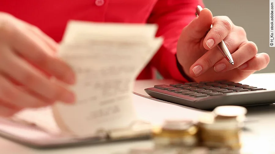 Person sitzt an einem Tisch und bearbeitet Rechnungen. Auf dem Tisch steht ein Sparschwein und ein Stapel Münzen.