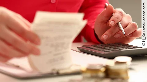 Rechnung Person sitzt an einem Tisch und bearbeitet Rechnungen. Auf dem Tisch steht ein Sparschwein und ein Stapel Münzen.