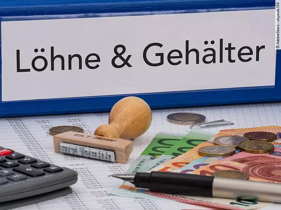 Ein dicker Ordner, auf dem "Löhne & Gehälter" steht. Davor liegen ein Taschenrechner, einige Geldscheine, ein Stempel und ein Kugelschreiber.
