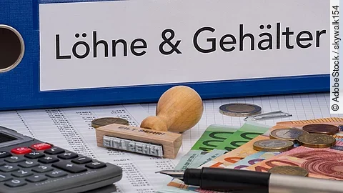 Ein dicker Ordner, auf dem "Löhne & Gehälter" steht. Davor liegen ein Taschenrechner, einige Geldscheine, ein Stempel und ein Kugelschreiber.