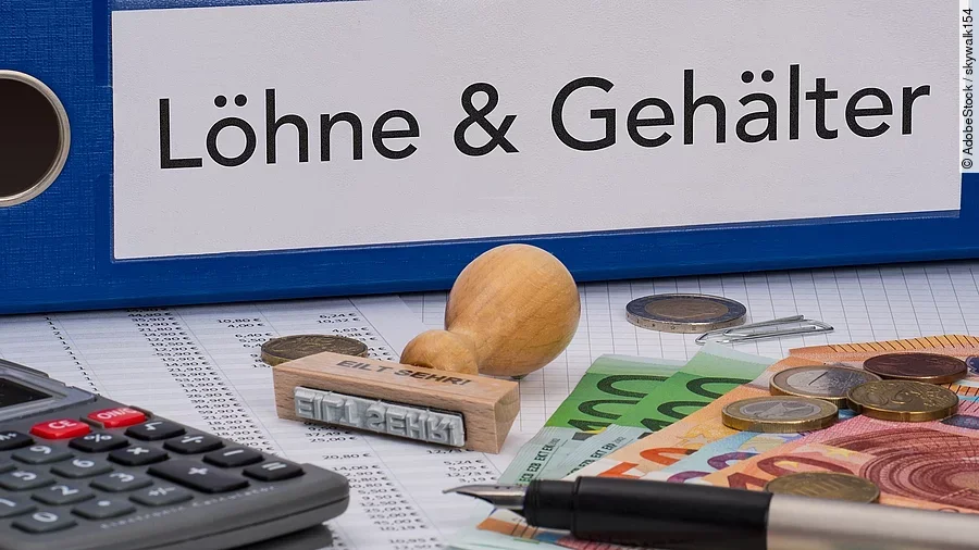 Ein dicker Ordner, auf dem "Löhne & Gehälter" steht. Davor liegen ein Taschenrechner, einige Geldscheine, ein Stempel und ein Kugelschreiber.
