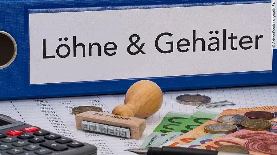 Ein dicker Ordner, auf dem "Löhne & Gehälter" steht. Davor liegen ein Taschenrechner, einige Geldscheine, ein Stempel und ein Kugelschreiber.