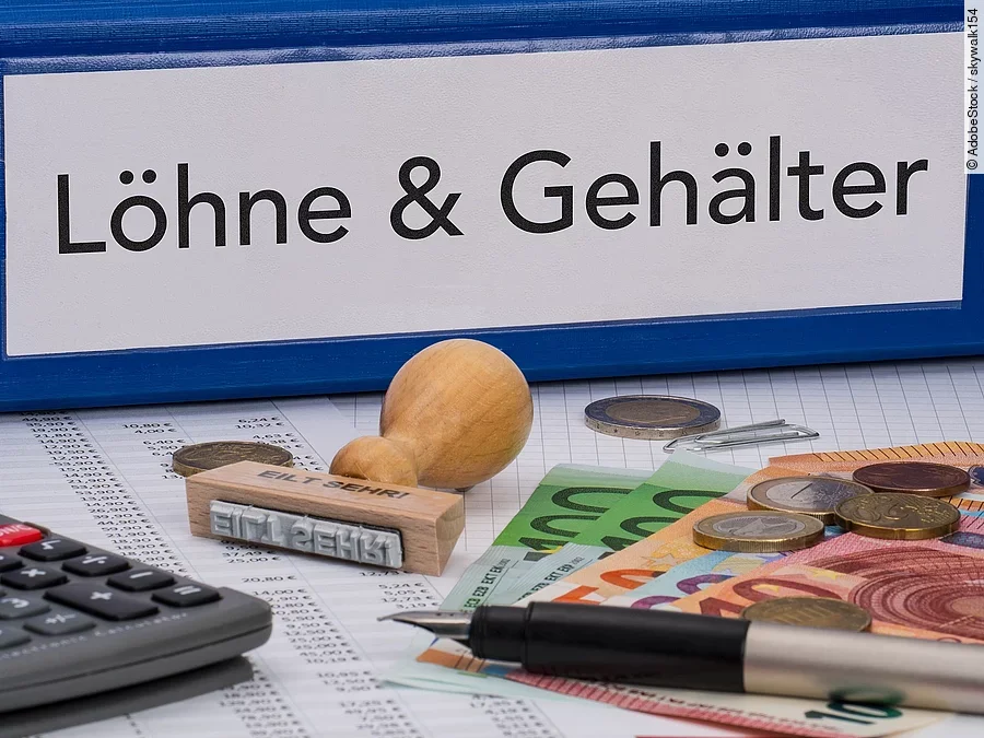 Ein dicker Ordner, auf dem "Löhne & Gehälter" steht. Davor liegen ein Taschenrechner, einige Geldscheine, ein Stempel und ein Kugelschreiber.