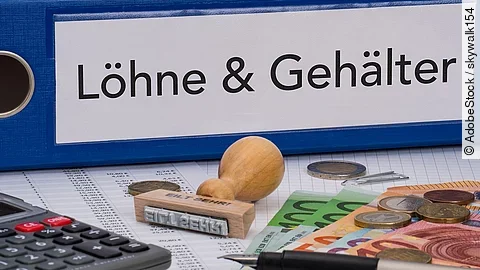 Ein dicker Ordner, auf dem "Löhne & Gehälter" steht. Davor liegen ein Taschenrechner, einige Geldscheine, ein Stempel und ein Kugelschreiber.