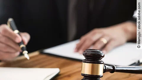 Ein Richterhammer liegt auf einem Tisch. Hinter im ist verschwommen ein Mann in Richterrobe zu sehen.