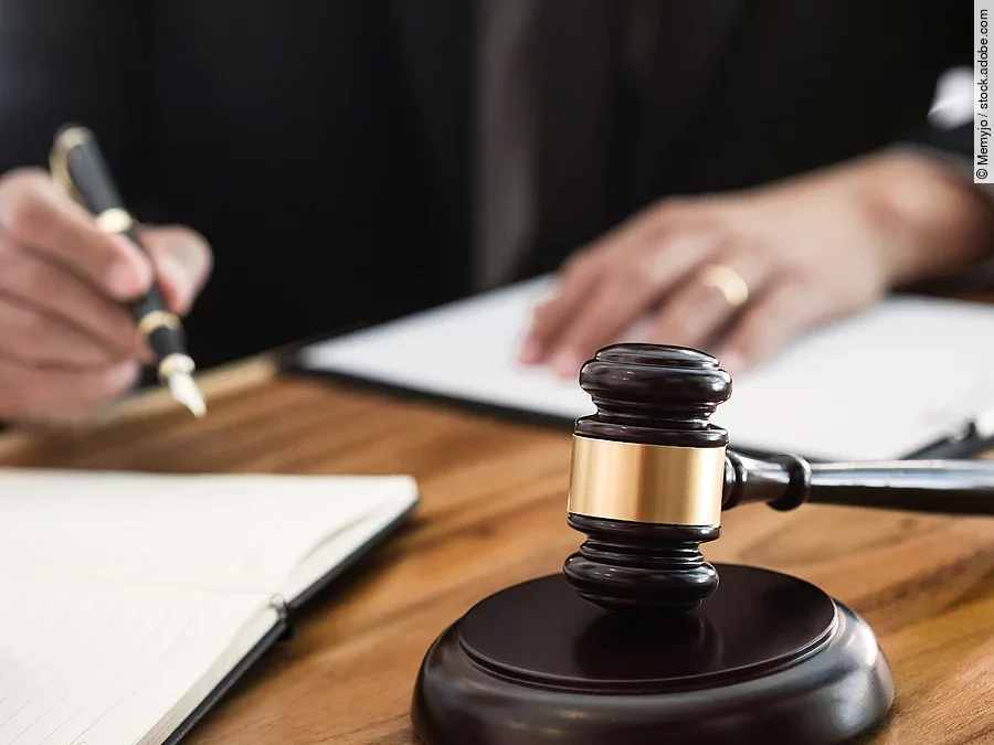 Ein Richterhammer liegt auf einem Tisch. Hinter im ist verschwommen ein Mann in Richterrobe zu sehen.