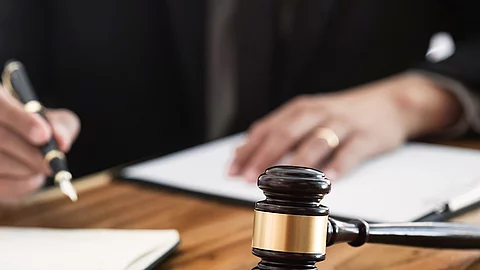 Ein Richterhammer liegt auf einem Tisch. Hinter im ist verschwommen ein Mann in Richterrobe zu sehen.