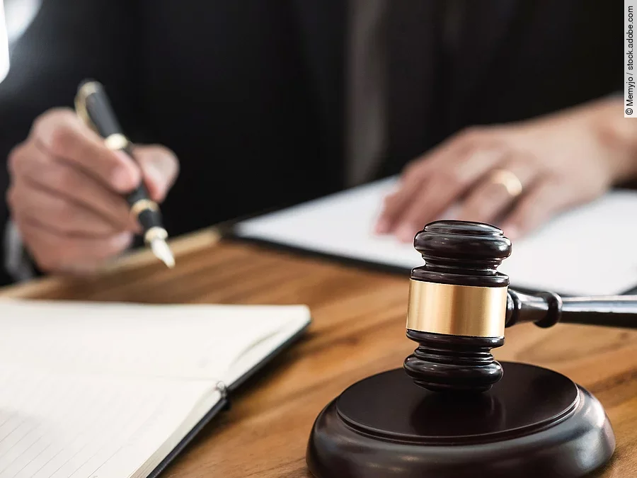 Ein Richterhammer liegt auf einem Tisch. Hinter im ist verschwommen ein Mann in Richterrobe zu sehen.