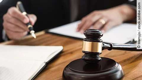 Rechtsspruch Ein Richterhammer liegt auf einem Tisch. Hinter im ist verschwommen ein Mann in Richterrobe zu sehen.