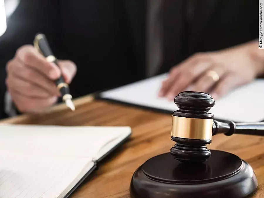 Ein Richterhammer liegt auf einem Tisch. Hinter im ist verschwommen ein Mann in Richterrobe zu sehen.