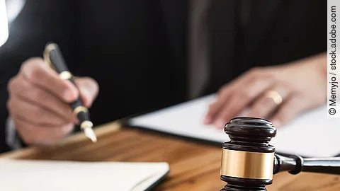 Ein Richterhammer liegt auf einem Tisch. Hinter im ist verschwommen ein Mann in Richterrobe zu sehen.