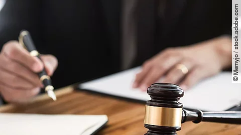 Ein Richterhammer liegt auf einem Tisch. Hinter im ist verschwommen ein Mann in Richterrobe zu sehen.