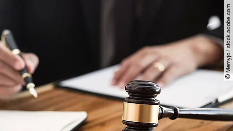 Ein Richterhammer liegt auf einem Tisch. Hinter im ist verschwommen ein Mann in Richterrobe zu sehen.