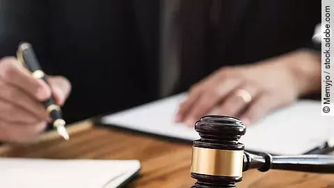 Ein Richterhammer liegt auf einem Tisch. Hinter im ist verschwommen ein Mann in Richterrobe zu sehen.