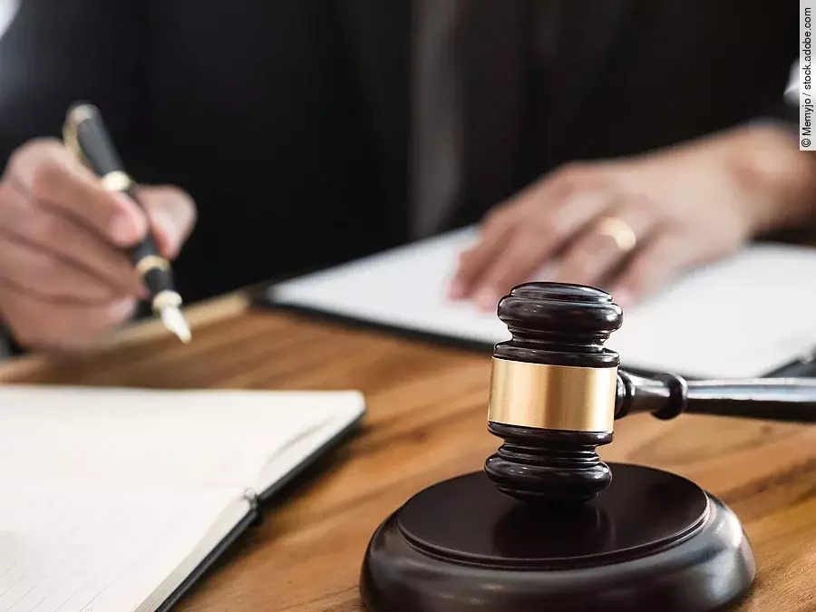 Ein Richterhammer liegt auf einem Tisch. Hinter im ist verschwommen ein Mann in Richterrobe zu sehen.