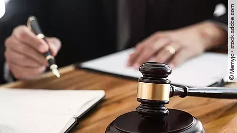 Ein Richterhammer liegt auf einem Tisch. Hinter im ist verschwommen ein Mann in Richterrobe zu sehen.