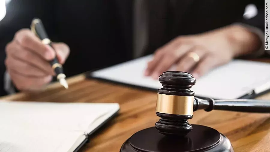 Ein Richterhammer liegt auf einem Tisch. Hinter im ist verschwommen ein Mann in Richterrobe zu sehen.