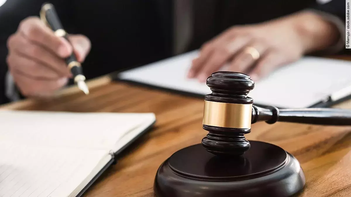 Rechtsspruch Ein Richterhammer liegt auf einem Tisch. Hinter im ist verschwommen ein Mann in Richterrobe zu sehen.