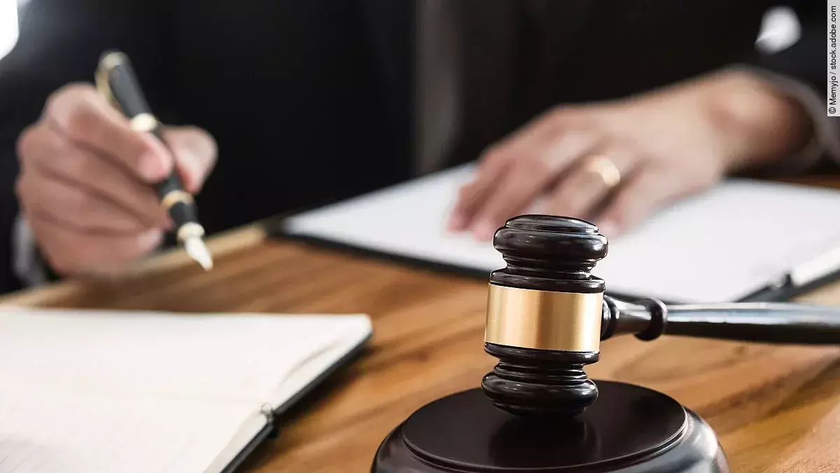 Ein Richterhammer liegt auf einem Tisch. Hinter im ist verschwommen ein Mann in Richterrobe zu sehen.