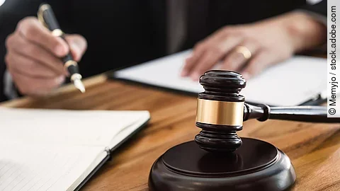 Ein Richterhammer liegt auf einem Tisch. Hinter im ist verschwommen ein Mann in Richterrobe zu sehen.