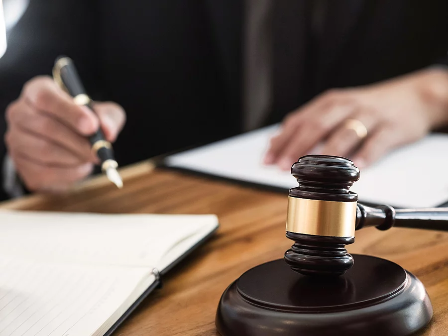 Ein Richterhammer liegt auf einem Tisch. Hinter im ist verschwommen ein Mann in Richterrobe zu sehen.