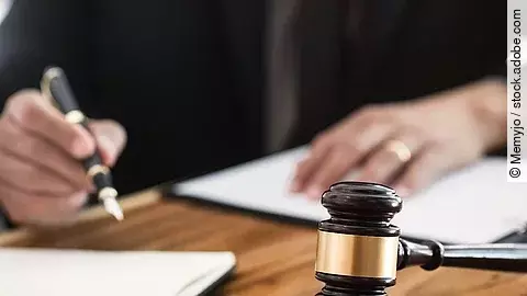 Ein Richterhammer liegt auf einem Tisch. Hinter im ist verschwommen ein Mann in Richterrobe zu sehen.