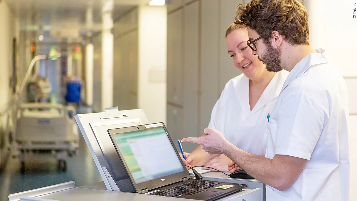 Eine Frau und ein Mann in weißen Kasacks schauen in ein Notebook, das auf dem Stationswagen im Krankenhausflur steht.