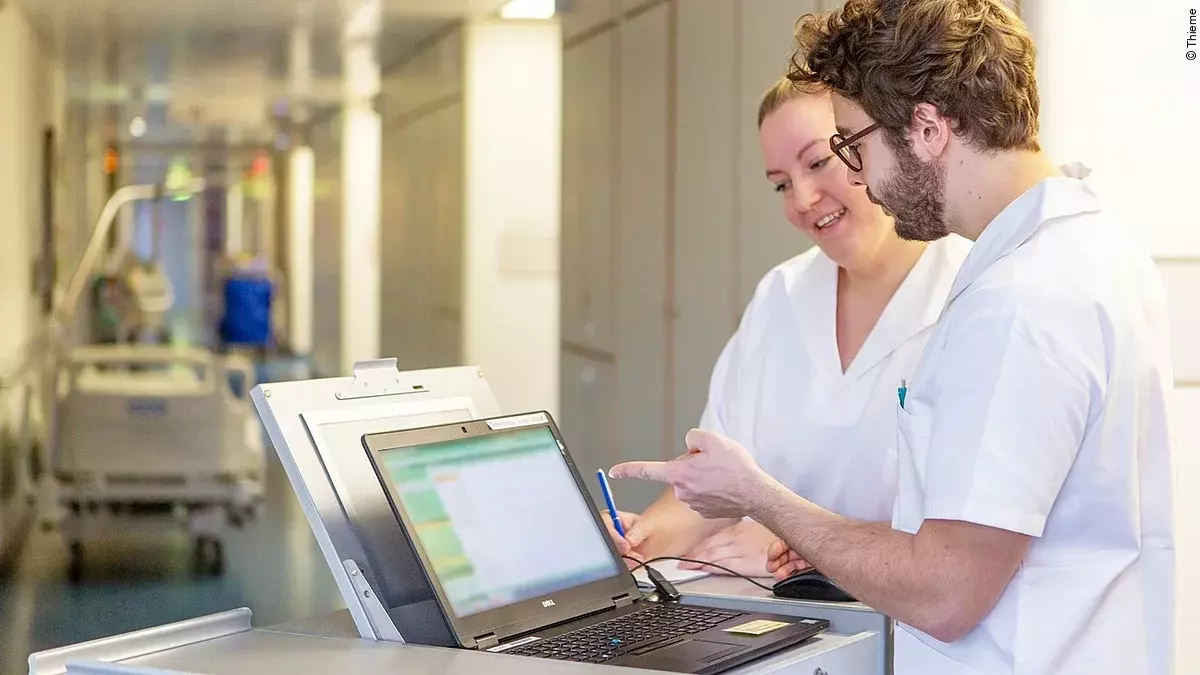 Eine Frau und ein Mann in weißen Kasacks schauen in ein Notebook, das auf dem Stationswagen im Krankenhausflur steht.