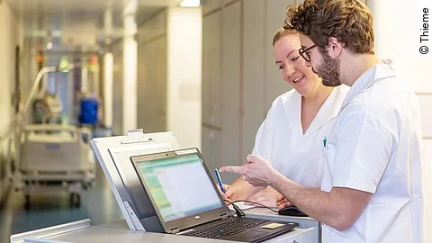 Eine Frau und ein Mann in weißen Kasacks schauen in ein Notebook, das auf dem Stationswagen im Krankenhausflur steht.