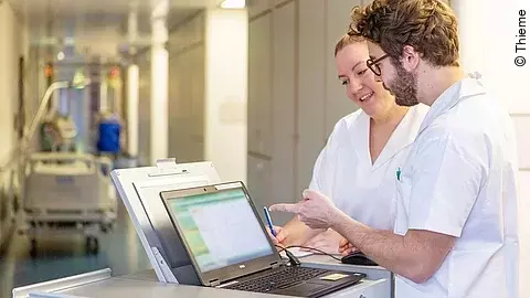 Pflegepersonal Eine Frau und ein Mann in weißen Kasacks schauen in ein Notebook, das auf dem Stationswagen im Krankenhausflur steht.