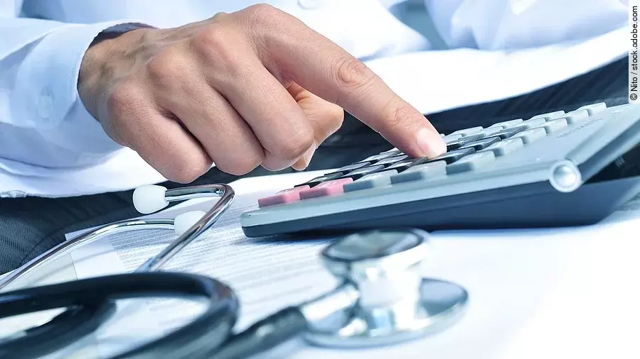 Die Hand eines Mannes in blauem Hemd tippt auf einem Taschenrechner. Daneben liegt ein Stethoskop.