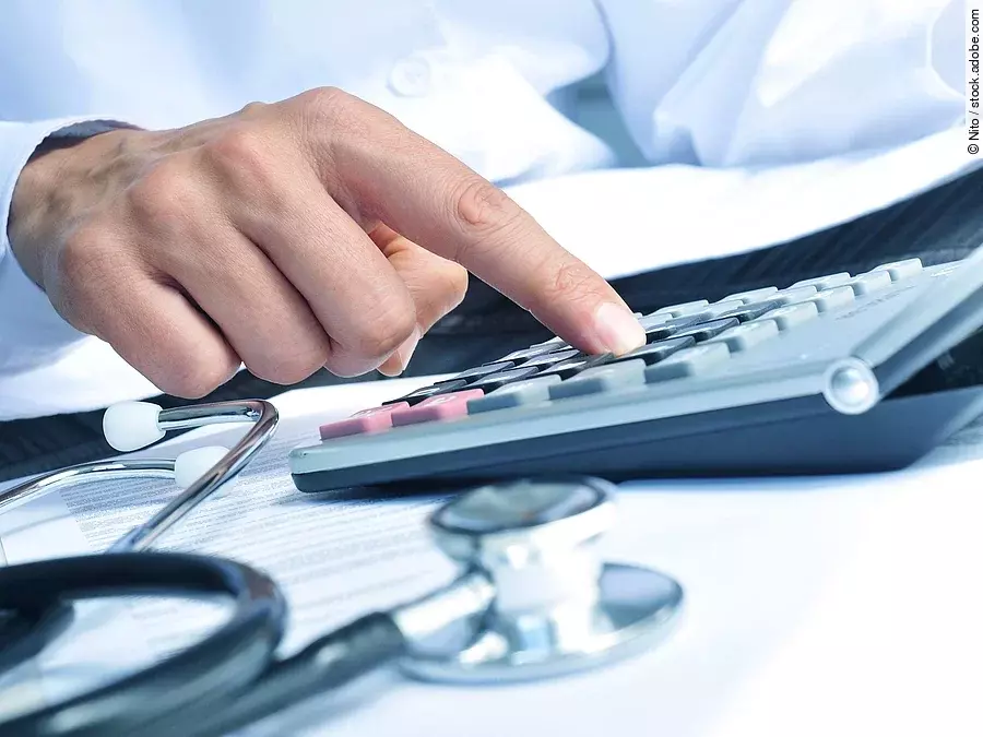 Die Hand eines Mannes in blauem Hemd tippt auf einem Taschenrechner. Daneben liegt ein Stethoskop.