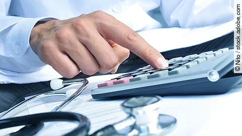 Die Hand eines Mannes in blauem Hemd tippt auf einem Taschenrechner. Daneben liegt ein Stethoskop.