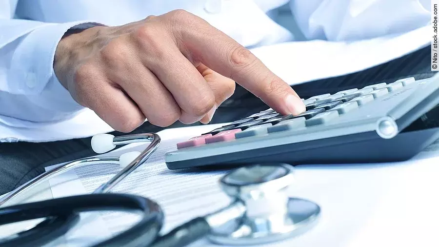 Die Hand eines Mannes in blauem Hemd tippt auf einem Taschenrechner. Daneben liegt ein Stethoskop.