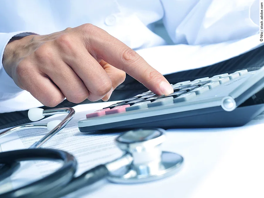 Taschenrechner Die Hand eines Mannes in blauem Hemd tippt auf einem Taschenrechner. Daneben liegt ein Stethoskop.
